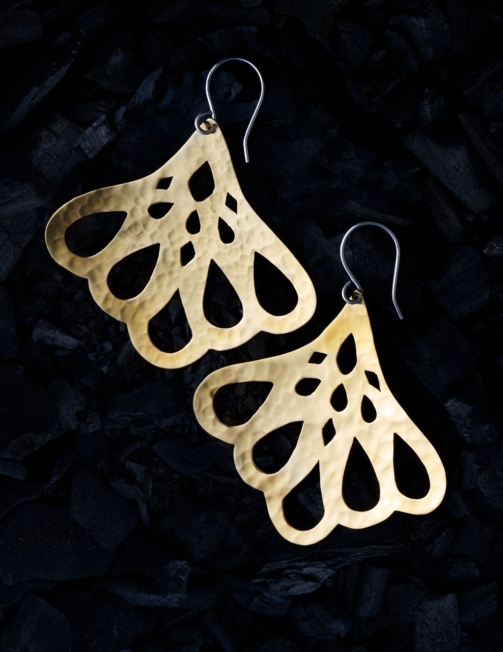 A pair of hammered gold-tone dangle earrings with an openwork scalloped design, displayed on a dark textured background of black charcoal.