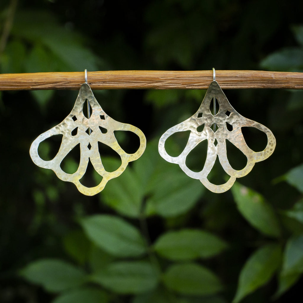 A pair of hammered silver-tone dangle earrings with an openwork floral pattern, hanging from a natural wooden branch against a blurred background of green leaves.