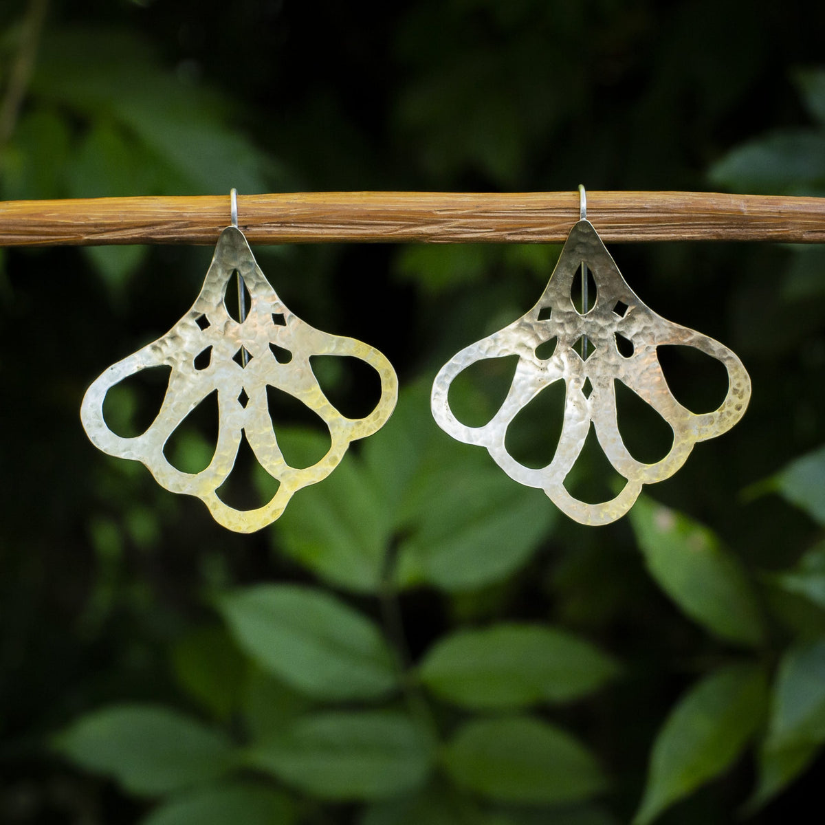 A pair of hammered silver-tone dangle earrings with an openwork floral pattern, hanging from a natural wooden branch against a blurred background of green leaves.