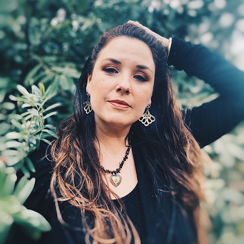 A woman with long dark brown hair posing outdoors in front of green foliage, wearing a black long-sleeve top, a black beaded necklace with a gold heart pendant, and gold dangling  lotus earrings.