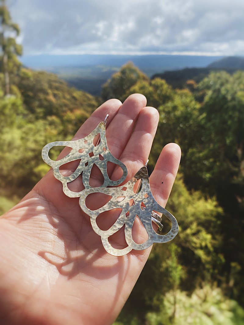 A pair of hammered silver-toned metal earrings with a butterfly wing silhouette, held in a person's hand against a blurred outdoor mountain and forest background.